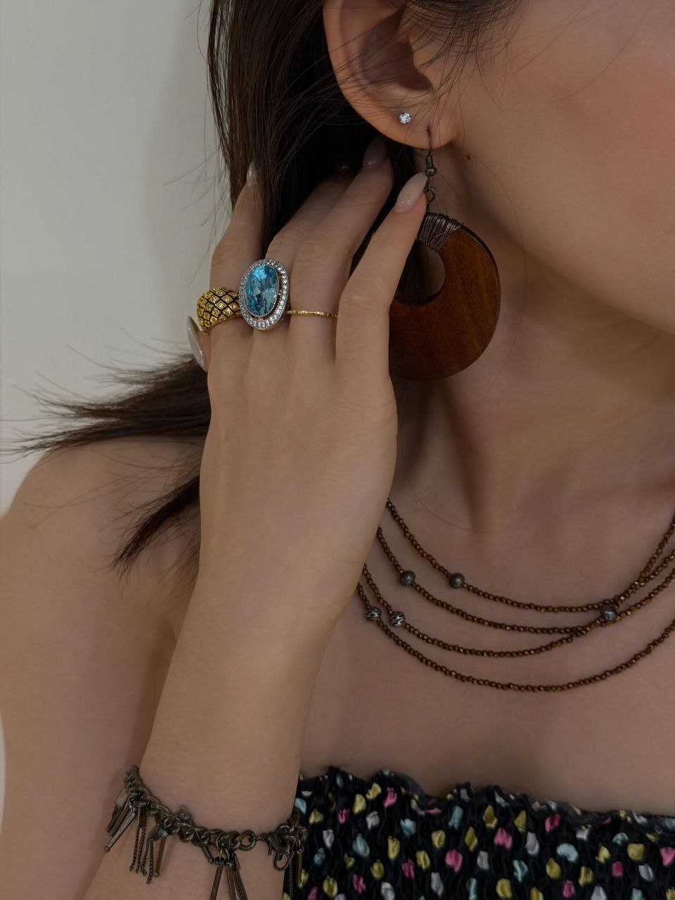 Hand detail showing blue topaz oval gemstone ring with diamond halo setting, paired with textured gold rings, and wooden drop earring visible