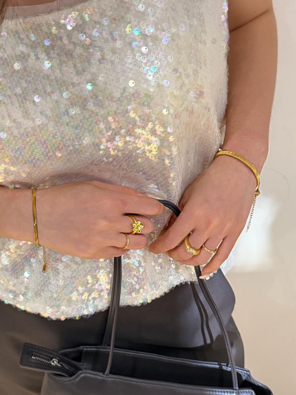 Hand detail holding black structured bag, showing stacked gold rings against luxury accessories and sequined top
