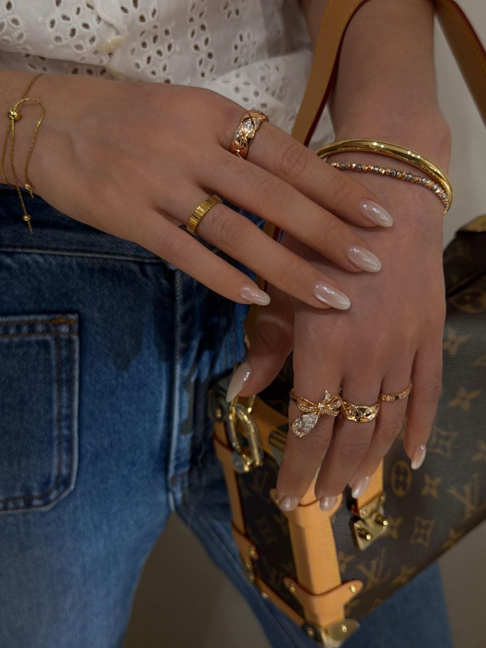 Hands wearing gold rings resting on blue jeans, with smooth gold bangle layered with beaded bracelet on wrist and delicate chain bracelet on opposite arm, monogram leather bag in background