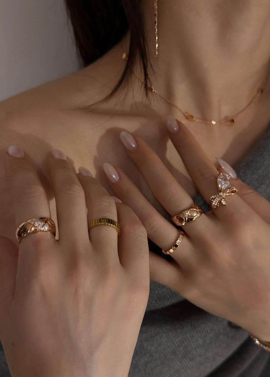 Two hands showing stacked gold rings including quilted textured rings, ribbed gold band, and bow-shaped ring with pear diamond, worn with gray knit top and layered gold necklace