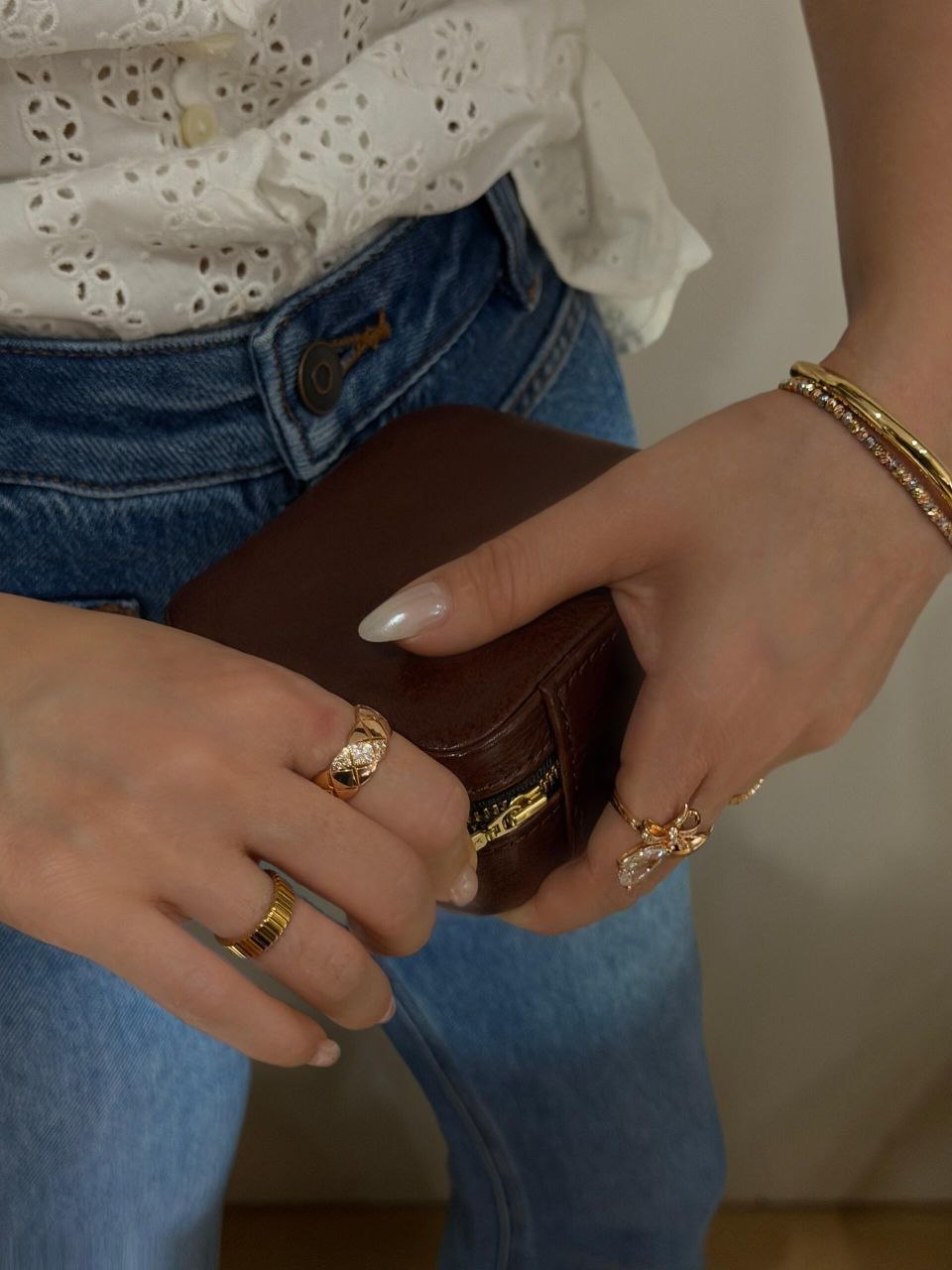 Hands with gold rings and layered gold bracelets holding small brown leather case, styled with white eyelet top and blue jeans