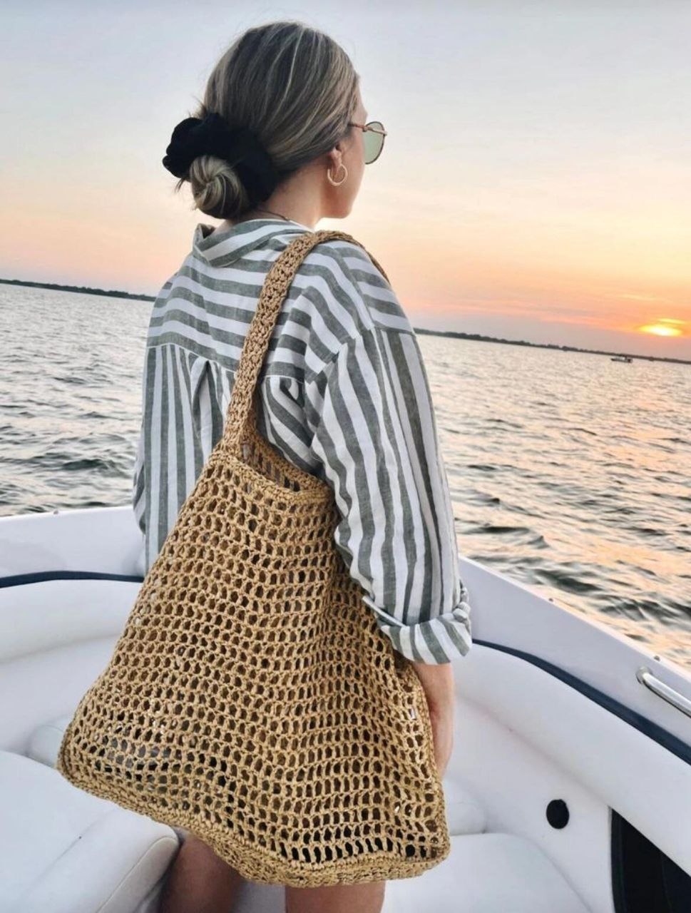 Back view of woman on boat at sunset wearing green striped shirt with open-weave crochet tote on shoulder, hair in low bun with black scrunchie