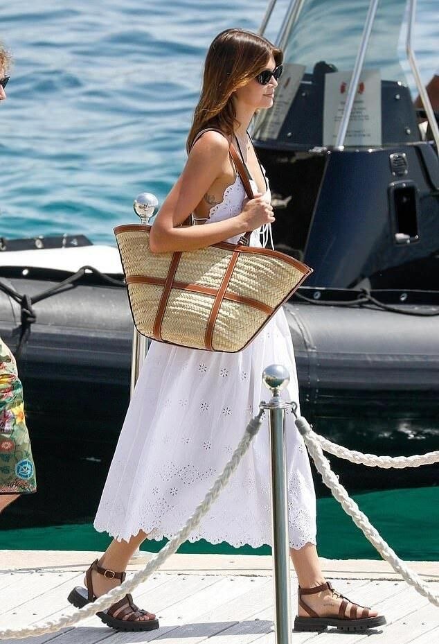Woman walking along dock in white eyelet midi dress carrying structured straw basket tote with brown leather trim, wearing brown fisherman sandals