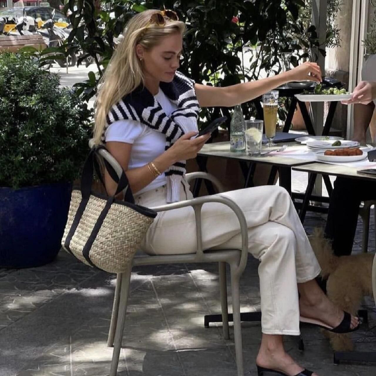 Woman at outdoor café in white tee and cream pants with navy striped sweater over shoulders, straw tote with black canvas straps beside her chair