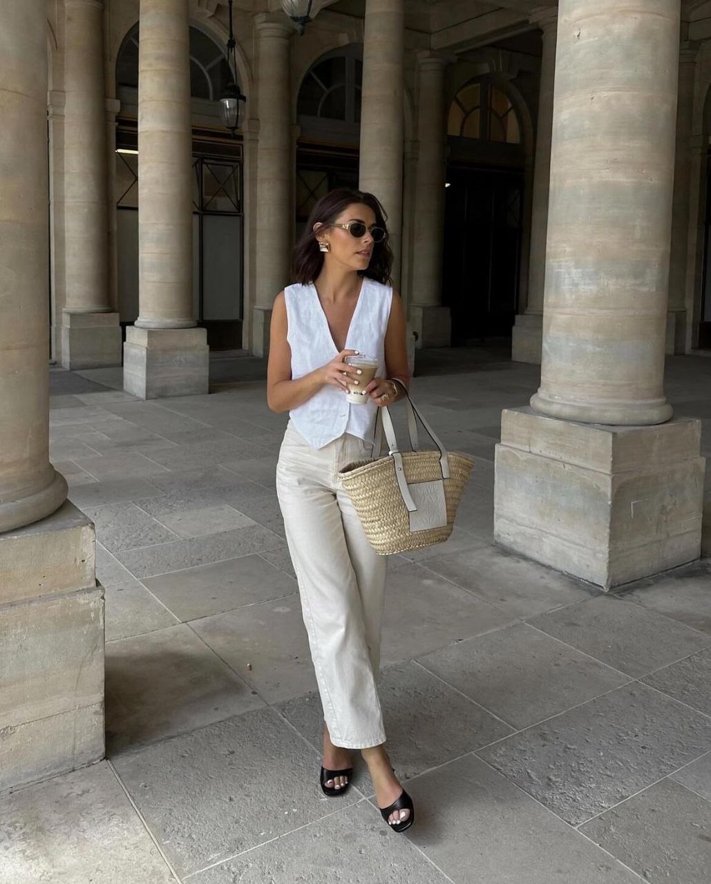 Woman in white linen vest and cream trousers with black slide sandals carrying straw basket tote with white leather trim among stone columns