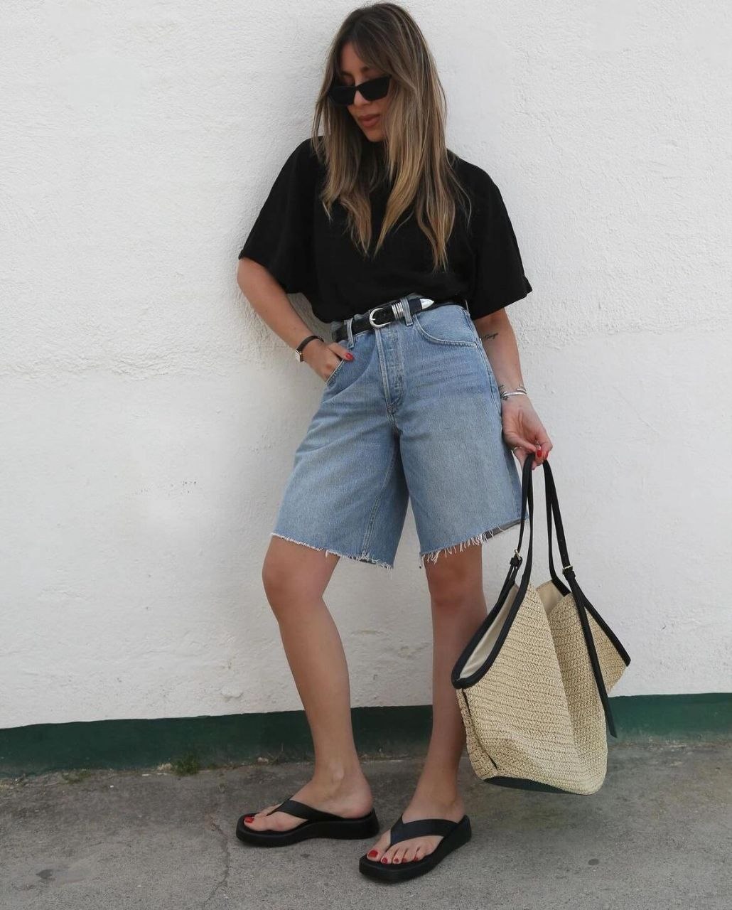 Woman leaning on white wall in black oversized tee and long denim bermuda shorts with western belt, carrying large soft woven tote with black straps