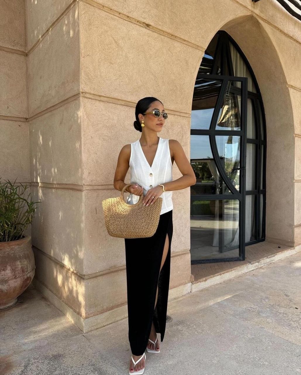 Woman in white button vest and black maxi skirt with high slit carrying round-handle woven handbag, styled with white thong heels and sunglasses