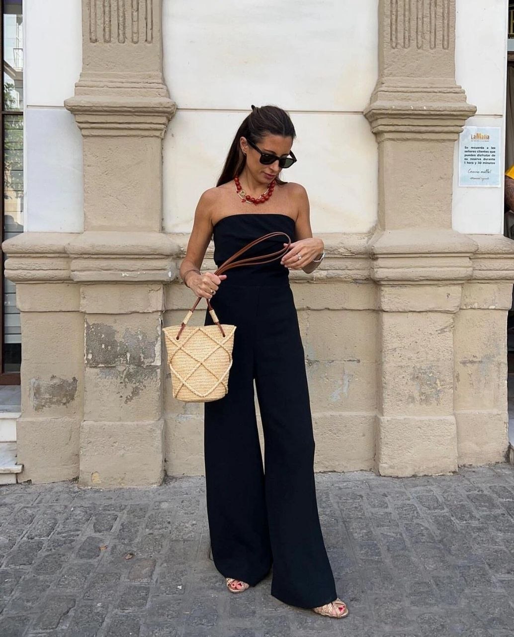 Woman in black strapless jumpsuit with red beaded necklace carrying small woven bucket bag with brown leather straps