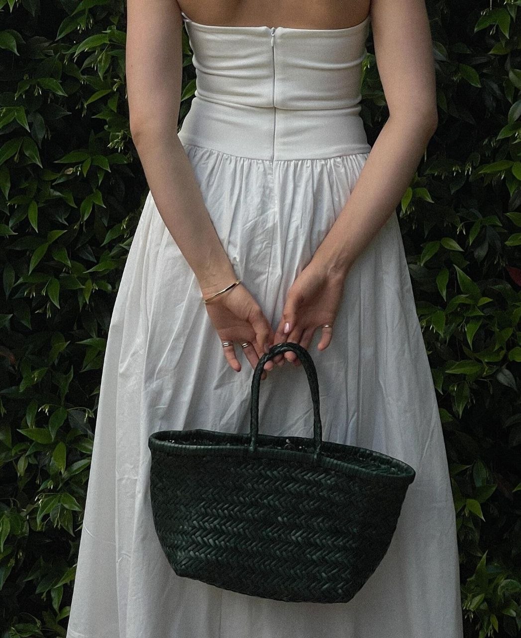 Back view of woman in white strapless drop-waist maxi dress holding small dark green woven leather basket bag against ivy backdrop