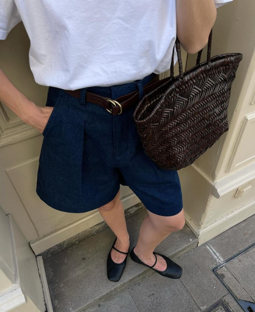 Detail of outfit with white tee, pleated dark denim shorts, layered brown leather belt, dark brown woven leather tote, and black mary jane flats