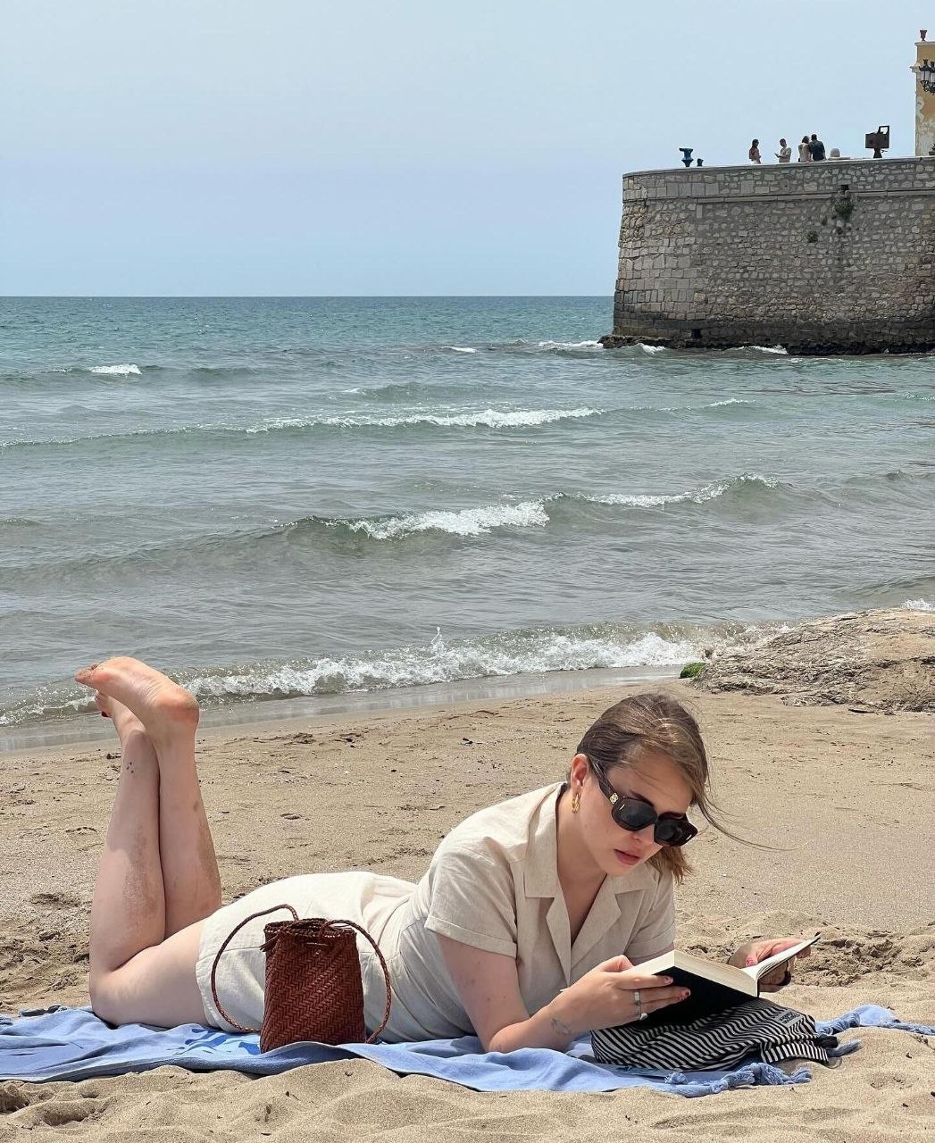 Woman reading a book on beach towel in cream linen set with small tan woven leather bucket bag beside her
