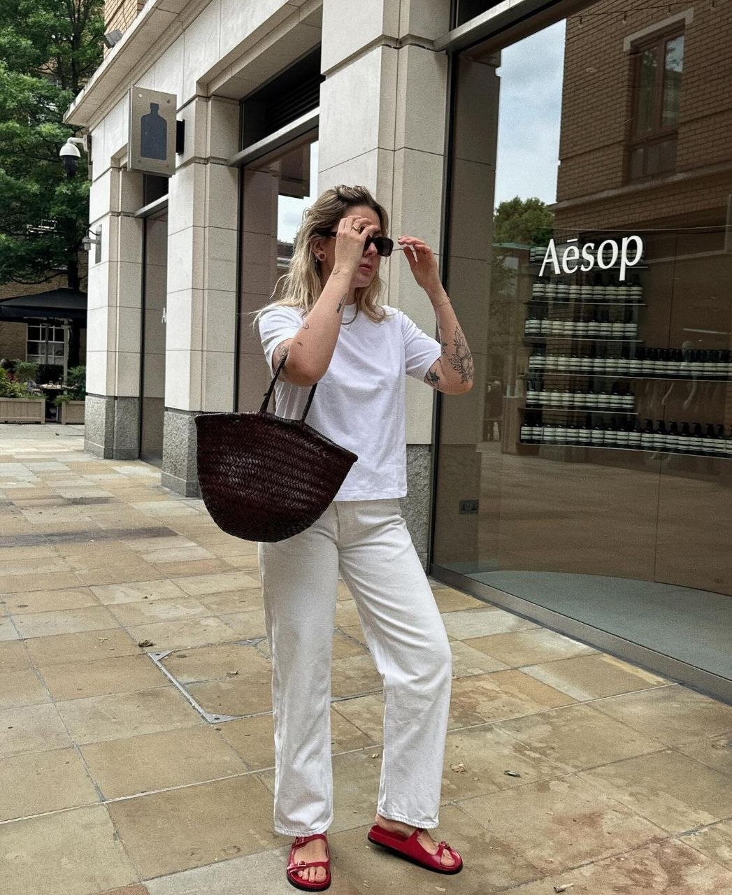 Woman in white tee and off-white jeans with red slide sandals carrying dark brown woven leather shoulder bag on city street