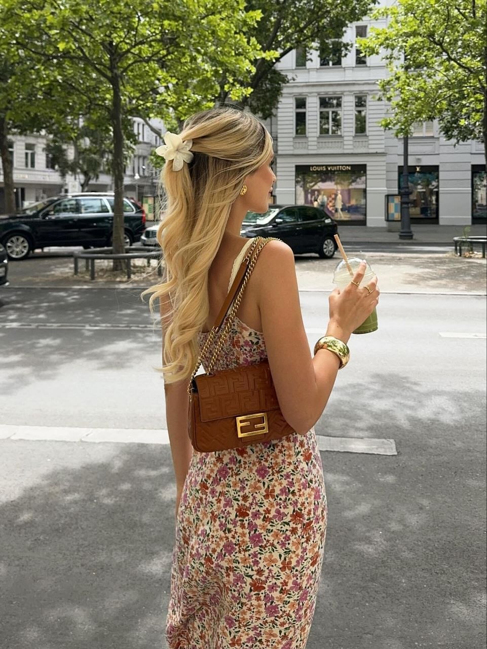 Woman with long blonde waves in half-up hairstyle secured by white flower clip, wearing floral midi dress with brown shoulder bag on city street