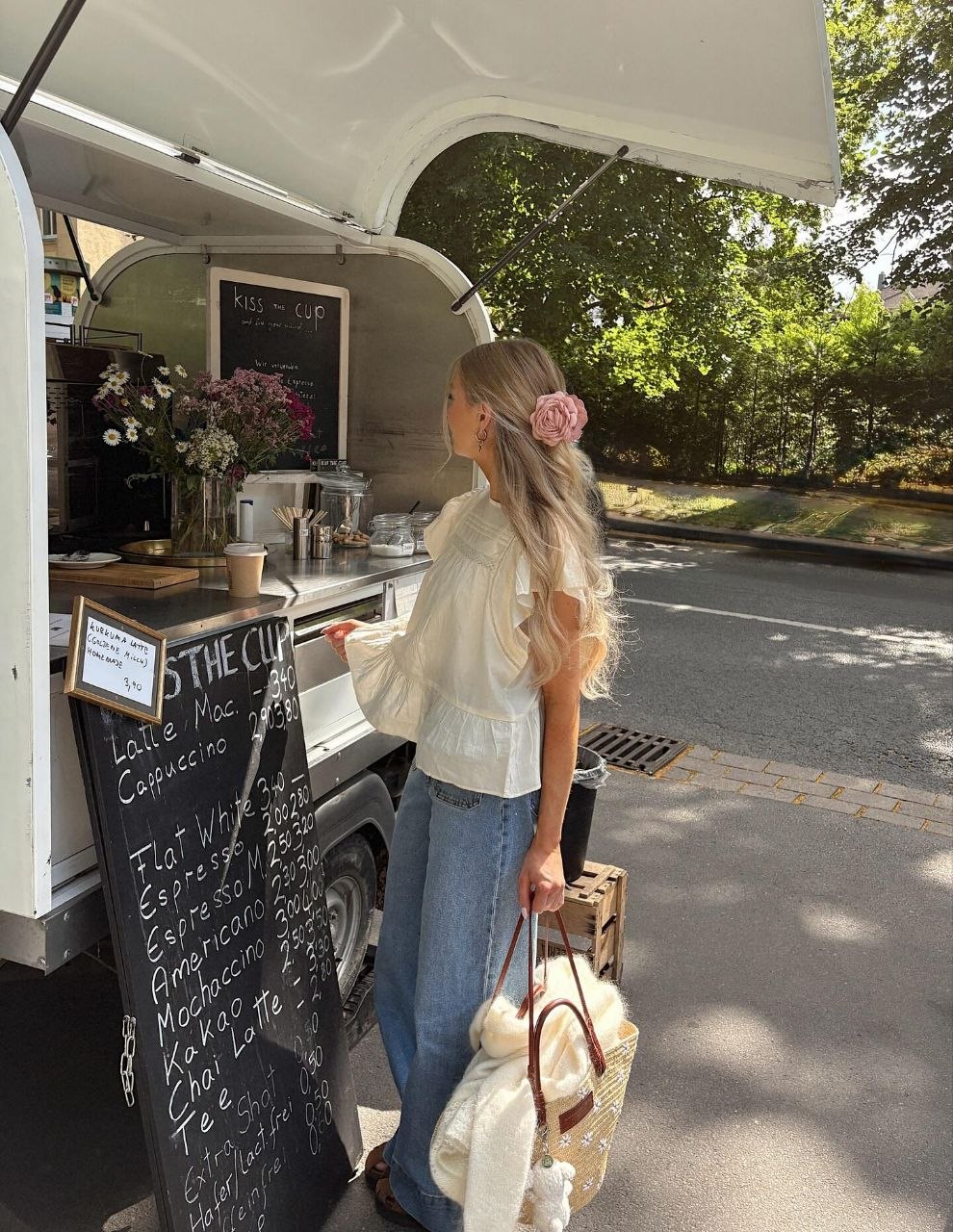 Woman at outdoor coffee cart wearing white ruffle blouse and wide-leg jeans with large pink rose clip in long wavy hair, carrying straw tote