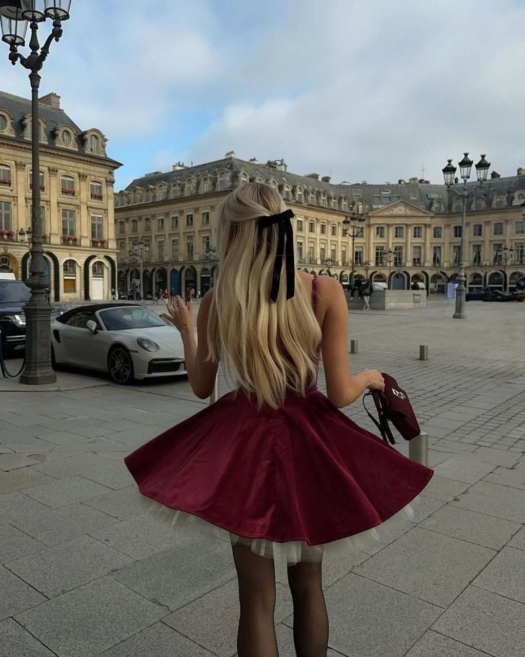 Back view of woman with long blonde hair tied in black velvet bow, wearing burgundy velvet mini dress with tulle underskirt in European square