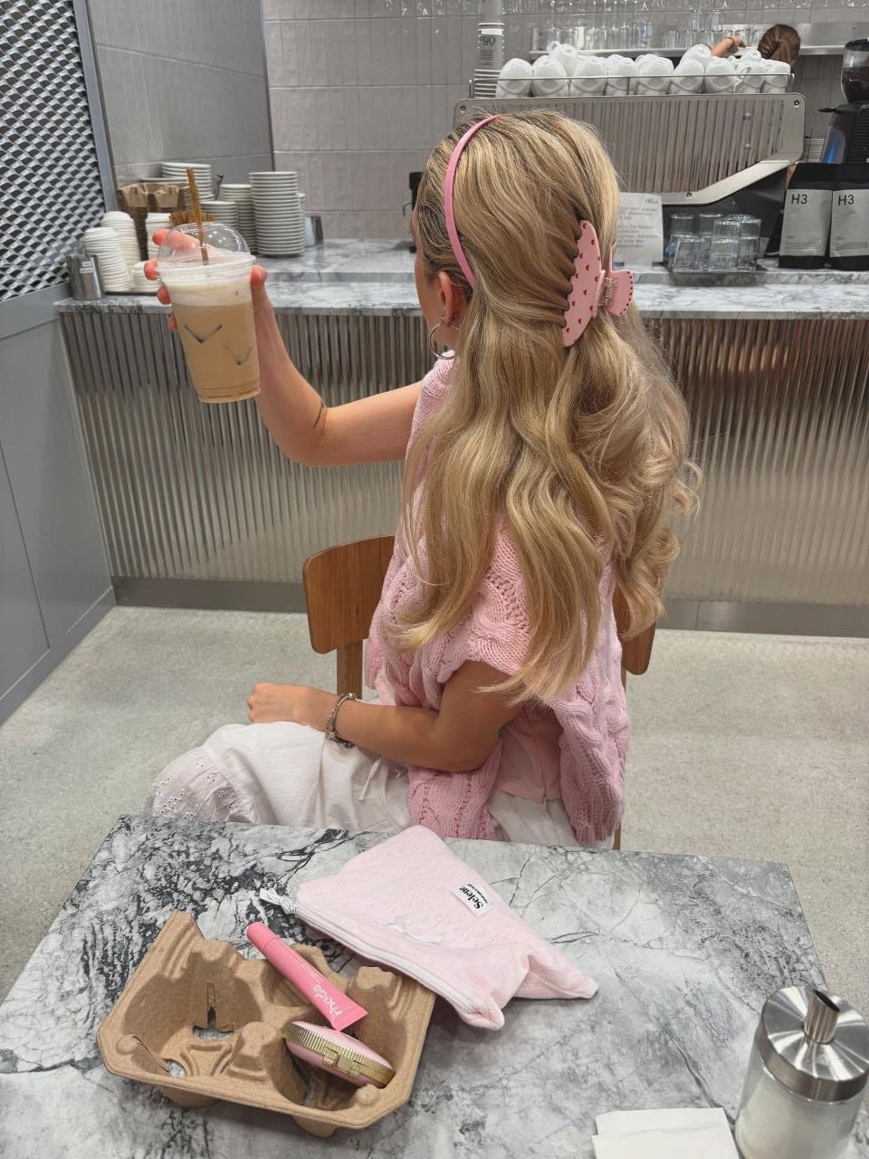 Woman in café wearing pink cable-knit vest with pink headband and heart-print claw clip holding half-up blonde waves