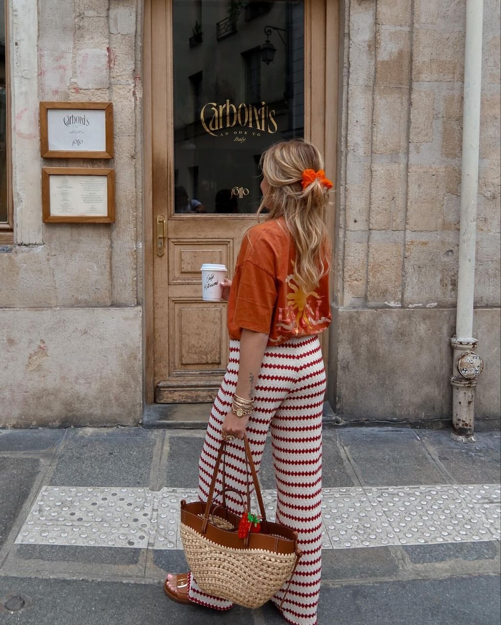 Woman from behind wearing orange graphic tee and red patterned knit pants with orange scrunchie in low ponytail, carrying woven basket tote