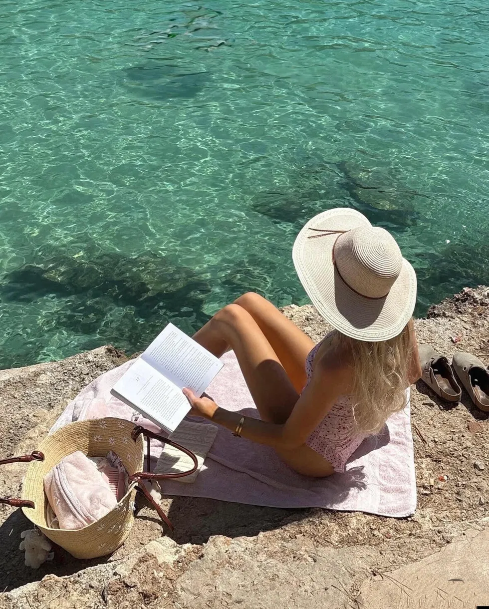 Woman wearing white wide-brim hat reading by the sea on a rocky beach