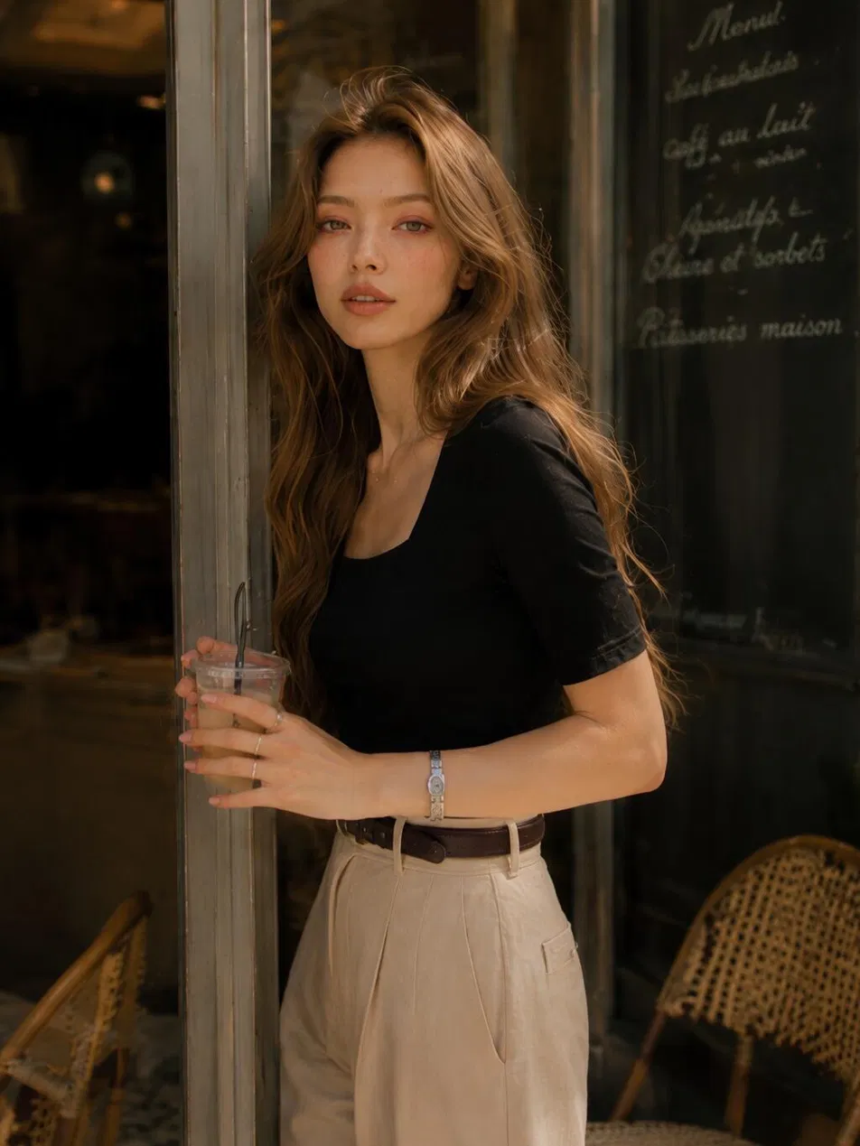 Woman in black tee tucked into neutral pleated pants with brown belt at cafe entrance