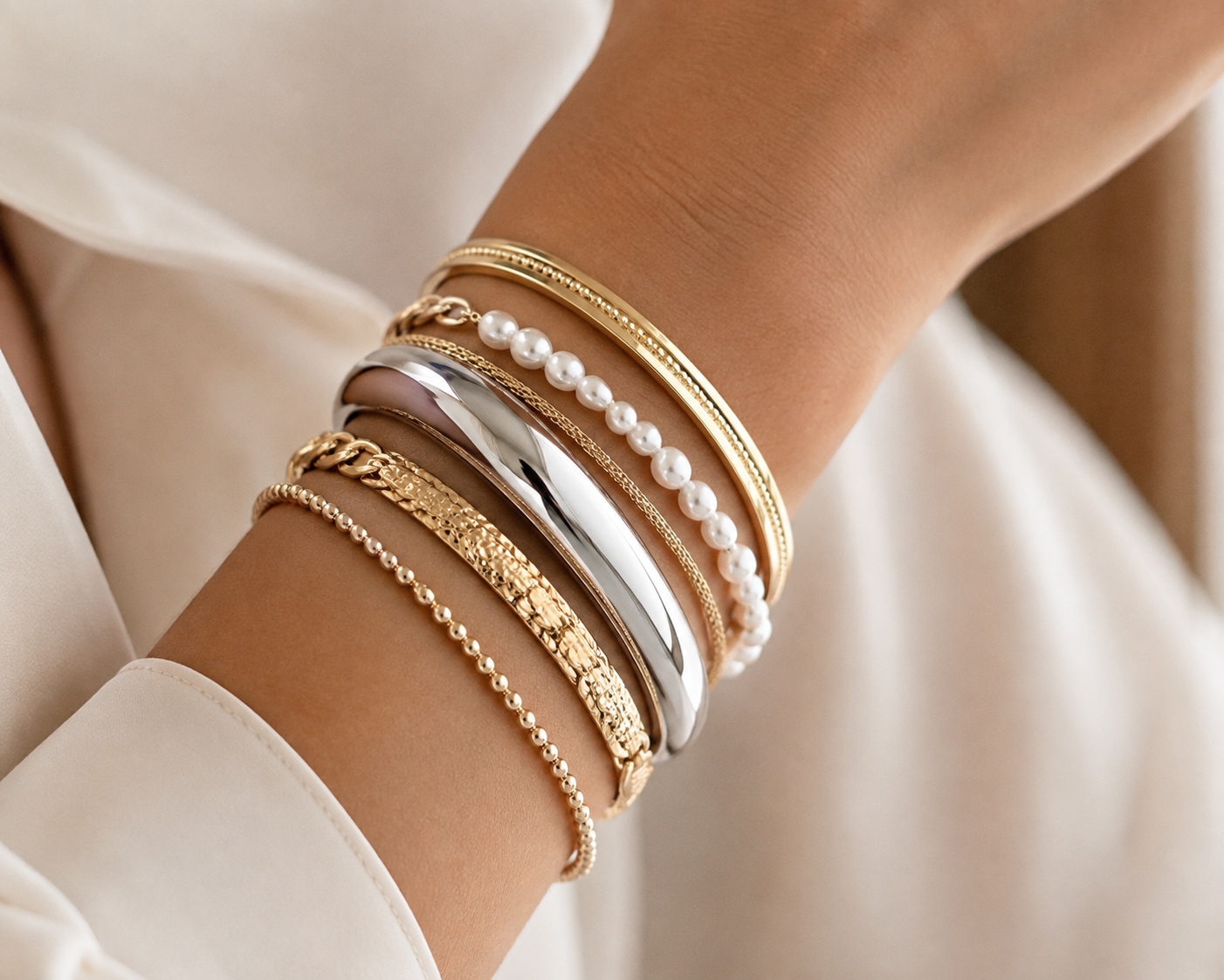 Woman's wrist wearing multiple stacked bracelets in different widths and materials (thin chains, chunky bangles, pearl bracelet), coordinated in metal tone