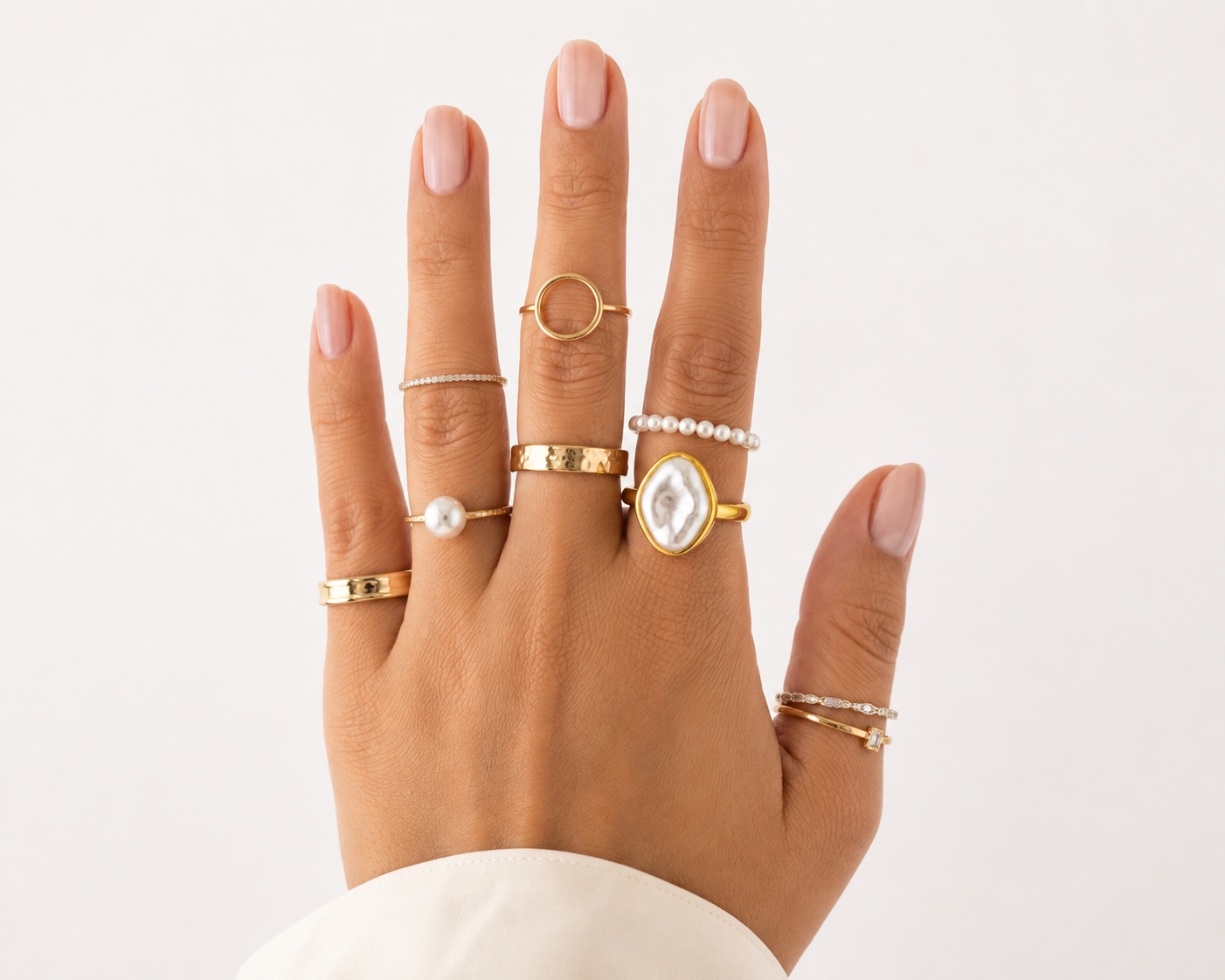 Woman's hand displaying multiple rings on different fingers (pearl ring, geometric rings, statement ring, thin bands), distributed across five fingers with coordinated metals