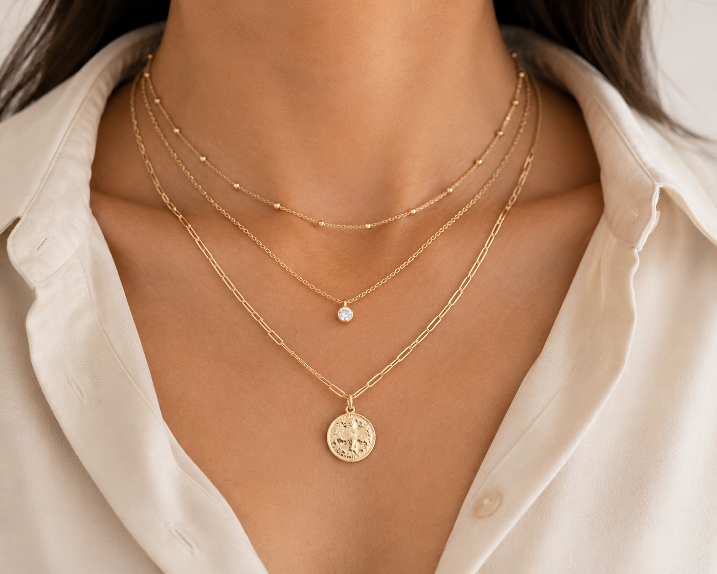 Woman's neck and chest showing three layered necklaces in varying lengths (short chain, medium pendant, long coin pendant), coordinated in warm gold tones