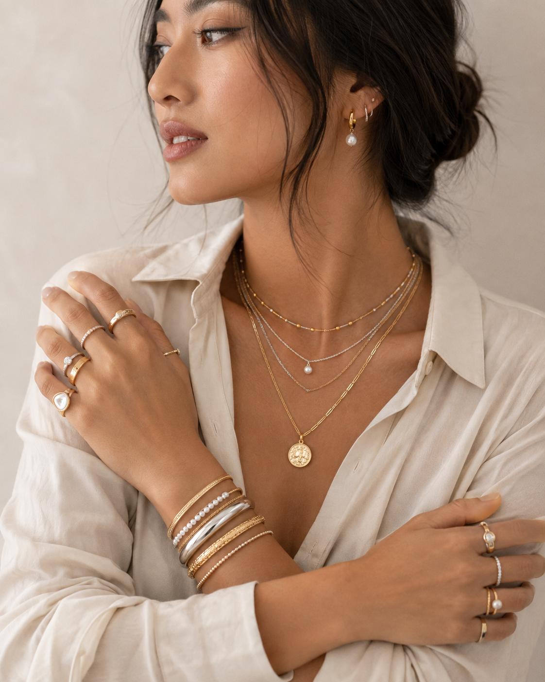 Woman wearing complete jewelry layering: stacked bracelets on wrist, multiple rings on fingers, layered necklaces, and pearl drop earrings, all in coordinated gold tones